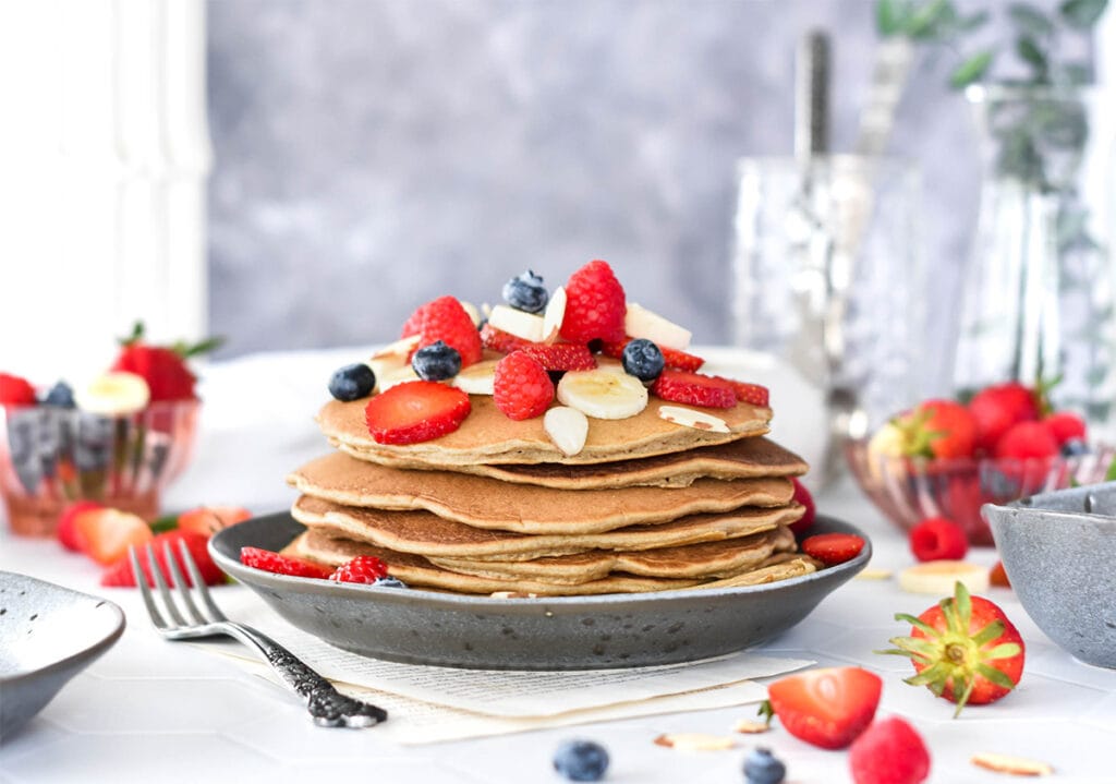 Stack of paleo banana flour pancakes topped with lots of banana, blueberries, strawberries and raspberries.