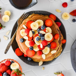 Stack of paleo pancakes topped with lots of banana, blueberries, strawberries and raspberries.