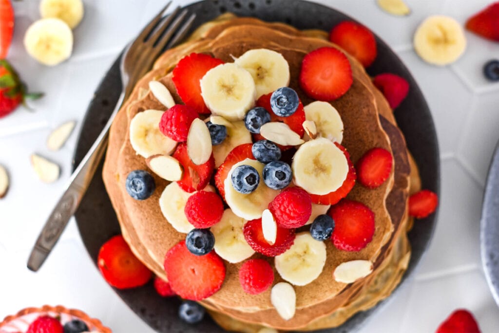 Stack of paleo banana flour pancakes topped with lots of banana, blueberries, strawberries and raspberries.