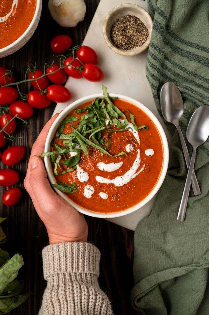 Hand holding a bowl of healthy paleo tomato soup topped with basil and coconut cream.