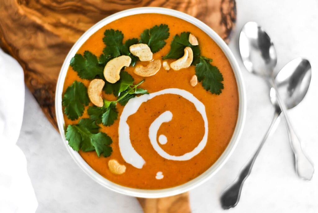 Bowl of bright orange curry butternut squash soup with cilantro, coconut cream and cashews on top.