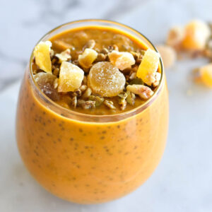 Glass filled with bright orange pumpkin chia pudding, topped with nuts and ginger.