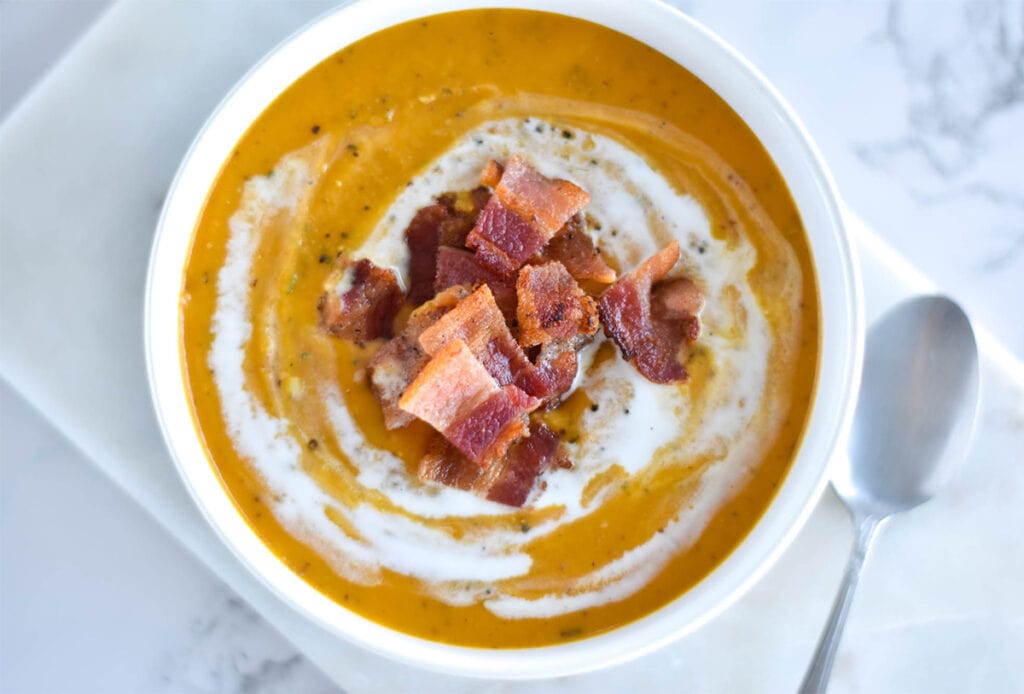 Thick bowl of orange soup topped with chunks of bacon and coconut cream.