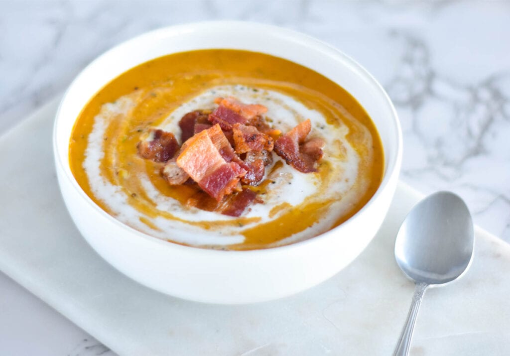 White bowl of orange sweet potato soup topped with a pile of bacon and coconut cream.