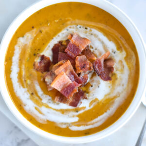 Thick bowl of orange soup topped with chunks of bacon and coconut cream.