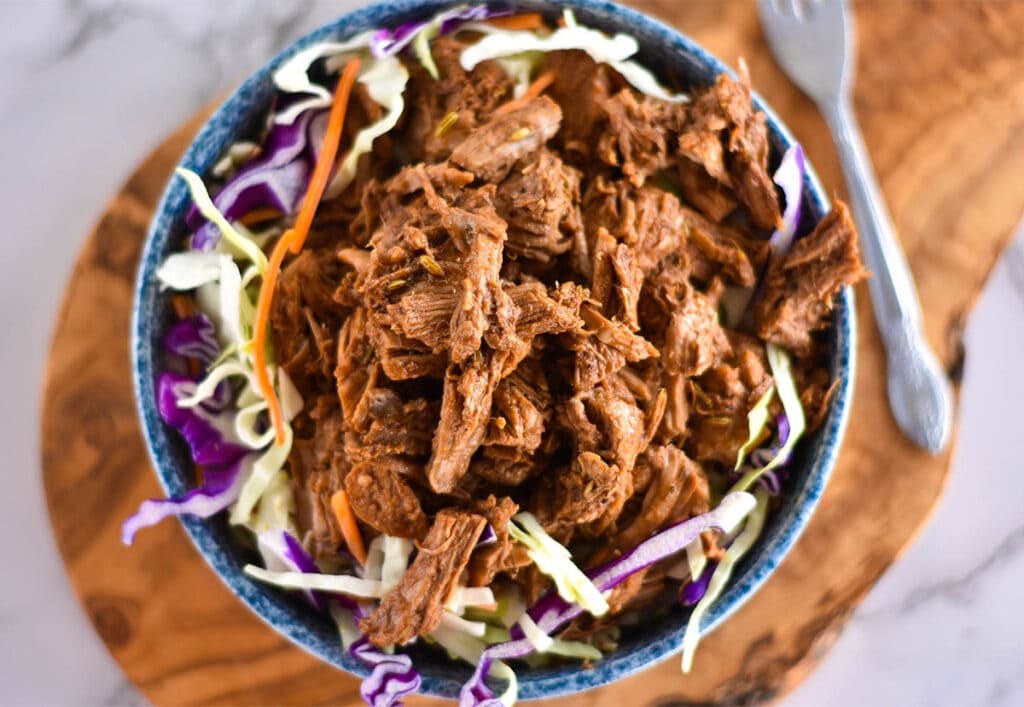 Top down view of blue bowl on a wooden cutting board, filled with coleslaw and pulled pork.