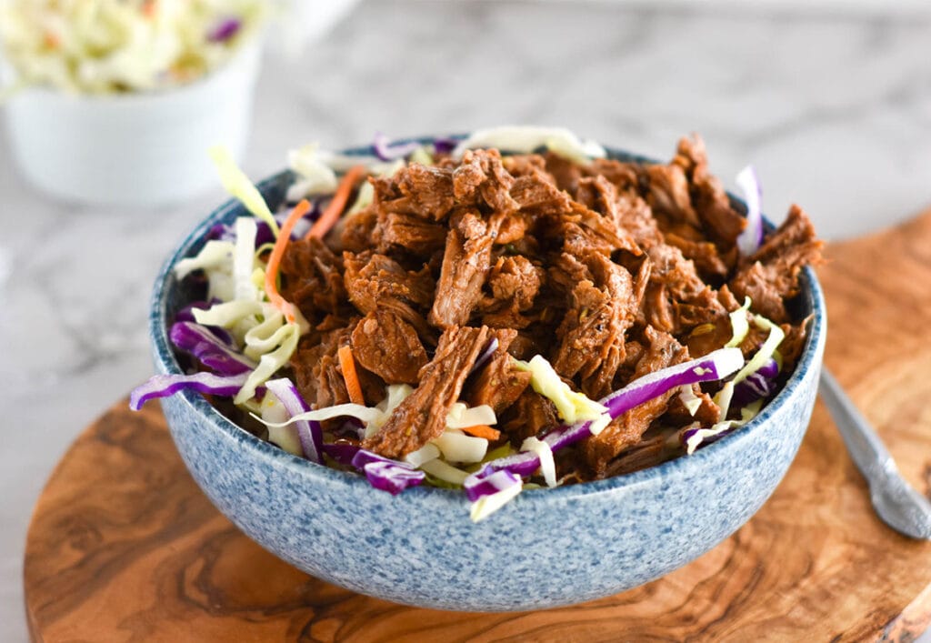 Blue bowl of pulled pork with coleslaw on a wooden serving board.