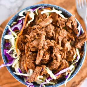 Top down view of blue bowl on a wooden cutting board, filled with coleslaw and pulled pork.