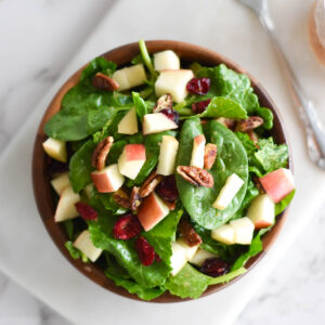 Wooden bowl full of spinach and kale with apples, pecans and cranberries next to a jar of dressing.