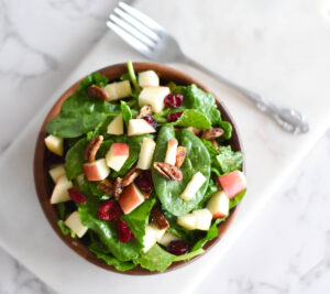 Wooden bowl full of spinach and kale with apples, pecans and cranberries next to a jar of dressing.