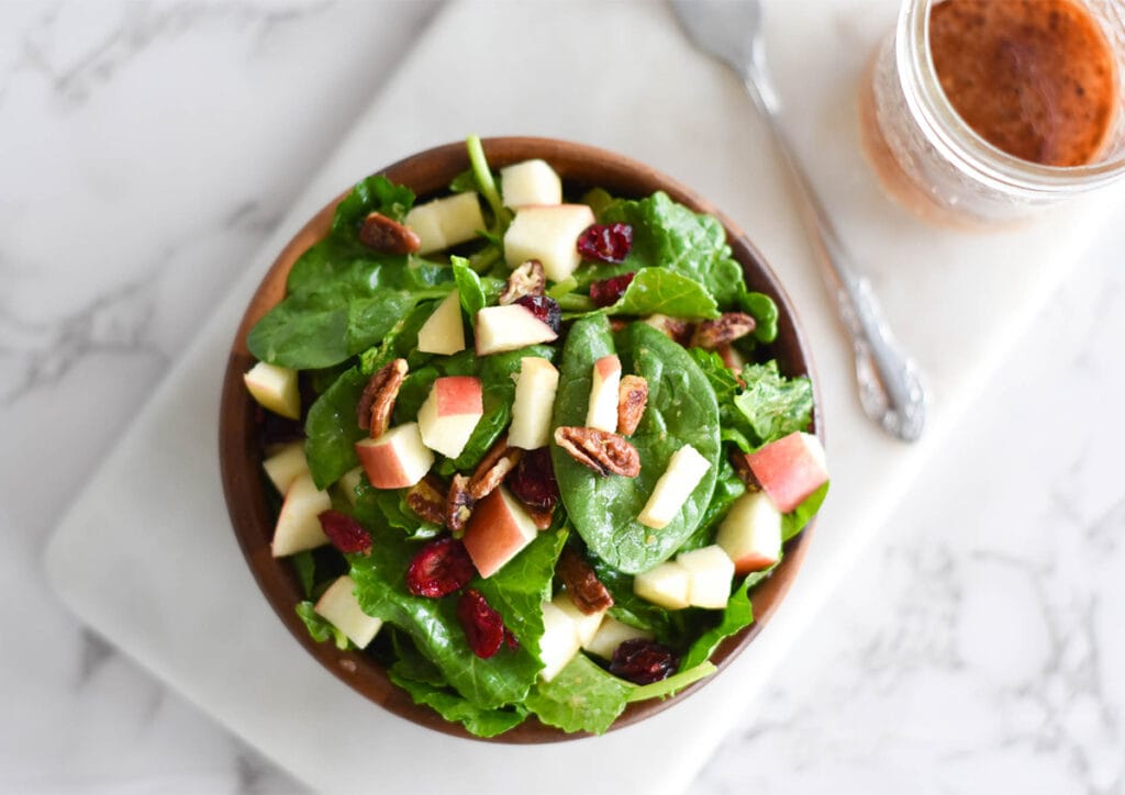 Wooden bowl full of spinach and kale with apples, pecans and cranberries next to a jar of dressing.