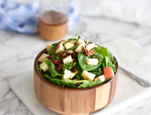 Wooden bowl on white marble serving board. Bowl is filled with green spinach, red apples, cranberries and pecans.