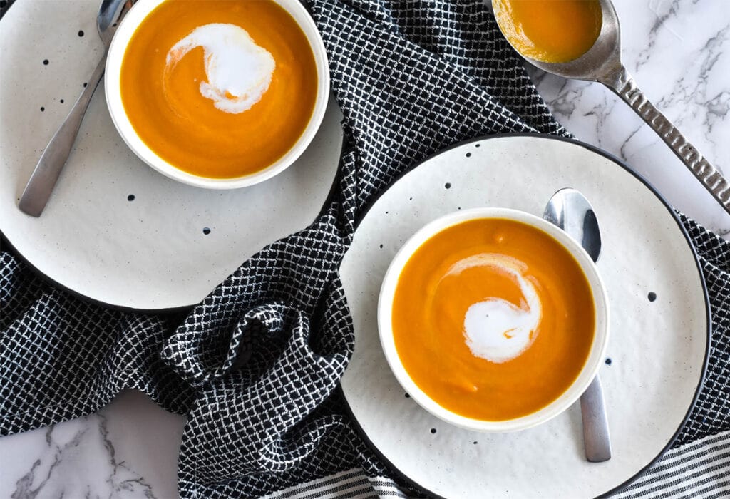 Two bowls of bright orange carrot ginger soup on white plates with spoons and a black towel.