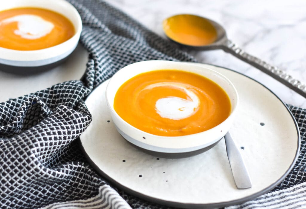 Side view of a blue, black and white bowl filled with carrot ginger soup. The soup is orange with a white coconut cream swirl.