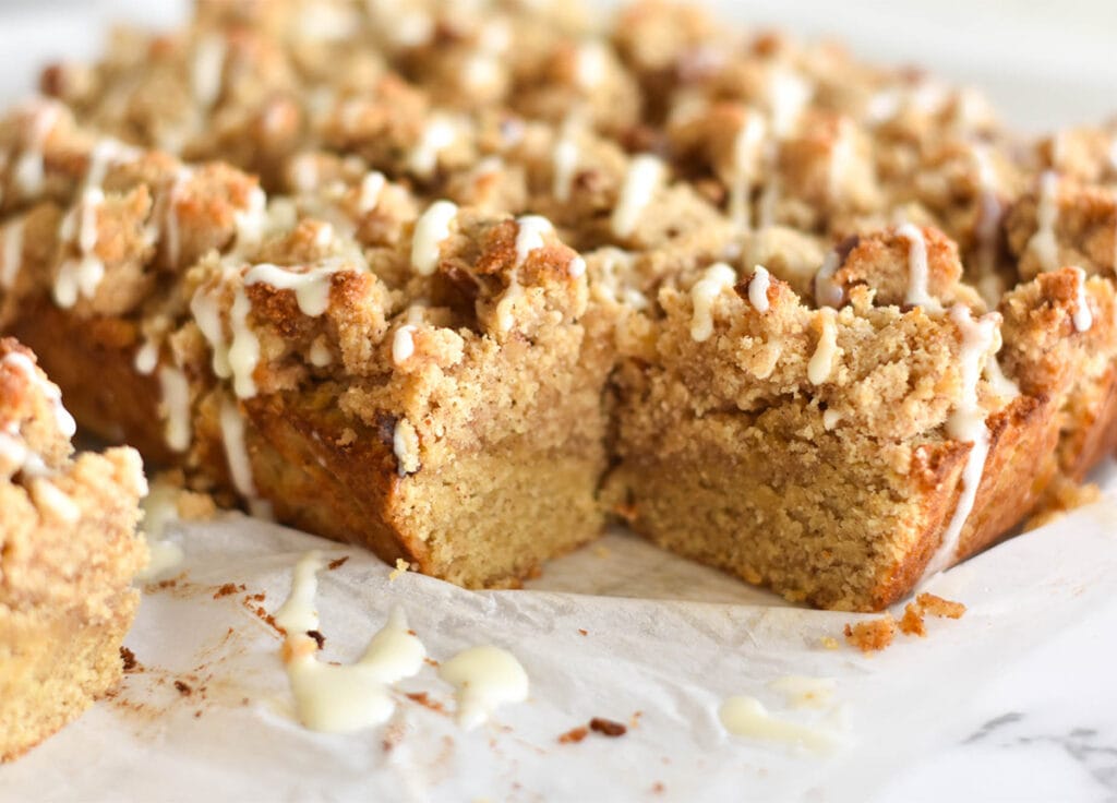 Keto cinnamon coffee cake with drizzled icing on parchment paper and one piece removed.