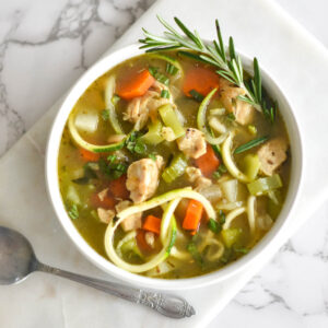 Paleo Chicken Zoodle Soup in a white bowl with carrots, potatoes and chicken breast, garnished with rosemary on a white cutting board.