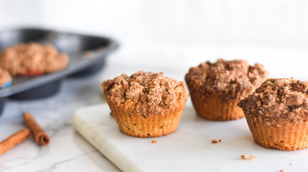 Paleo Coffee Cake Muffins on a white marble cutting board with a muffin tin and cinnamon sticks in the background.