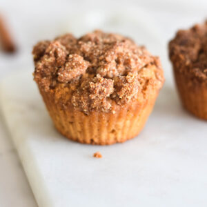 Close up on coffee cake muffin on white marble cutting board with cinnamon sticks in background.