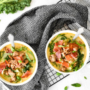 Two white bowls wrapped with a black towel filled with kale, sausage, potatoes, basil, green onion and a creamy yellow broth.