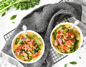 Two white bowls wrapped with a black towel filled with kale, sausage, potatoes, basil, green onion and a creamy yellow broth.