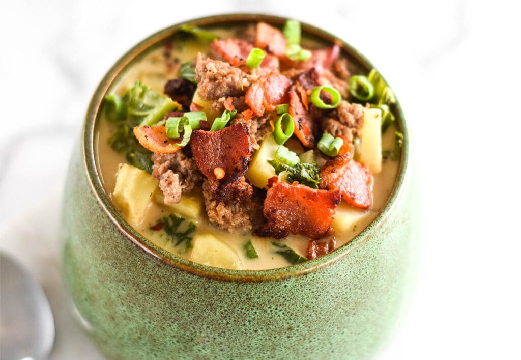 Green bowl filled to the brim with soup with kale, sausage, bacon, potatoes and chives.