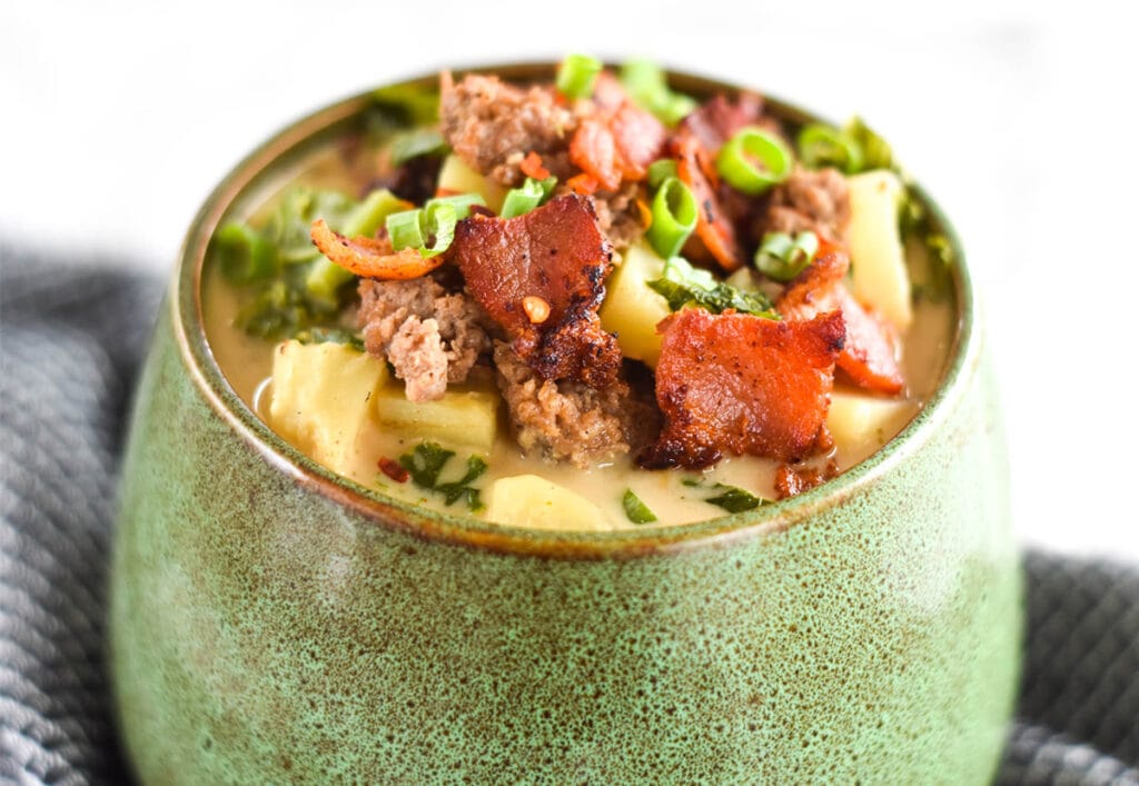 Green bowl filled to the brim with soup with zuppa toscana containing kale, sausage, bacon, potatoes and chives.