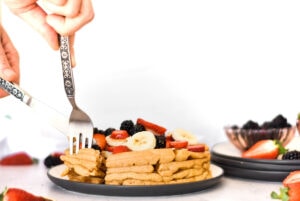 Fork and knife cutting into paleo waffles covered in bananas, blackberries, strawberries and almonds.