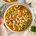 Hand touching a bowl of Paleo Chicken Noodle Soup with zucchini noodles.