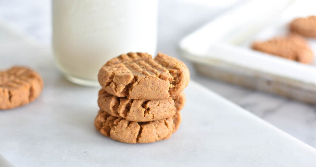 Stack of three keto peanut butter cookies next to tray of cookies and a glass of milk.