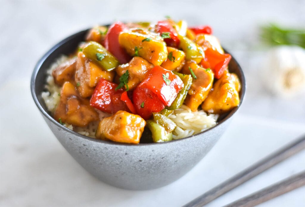 Bowl of paleo honey garlic chicken with red and green bell peppers on a bed of rice.