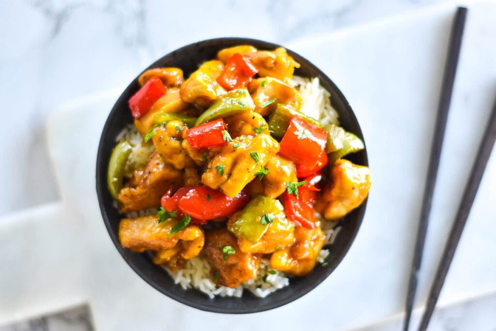 Close up shot of a dark bowl with brightly colored chicken and bell peppers on a bed of rice next to chop sticks.