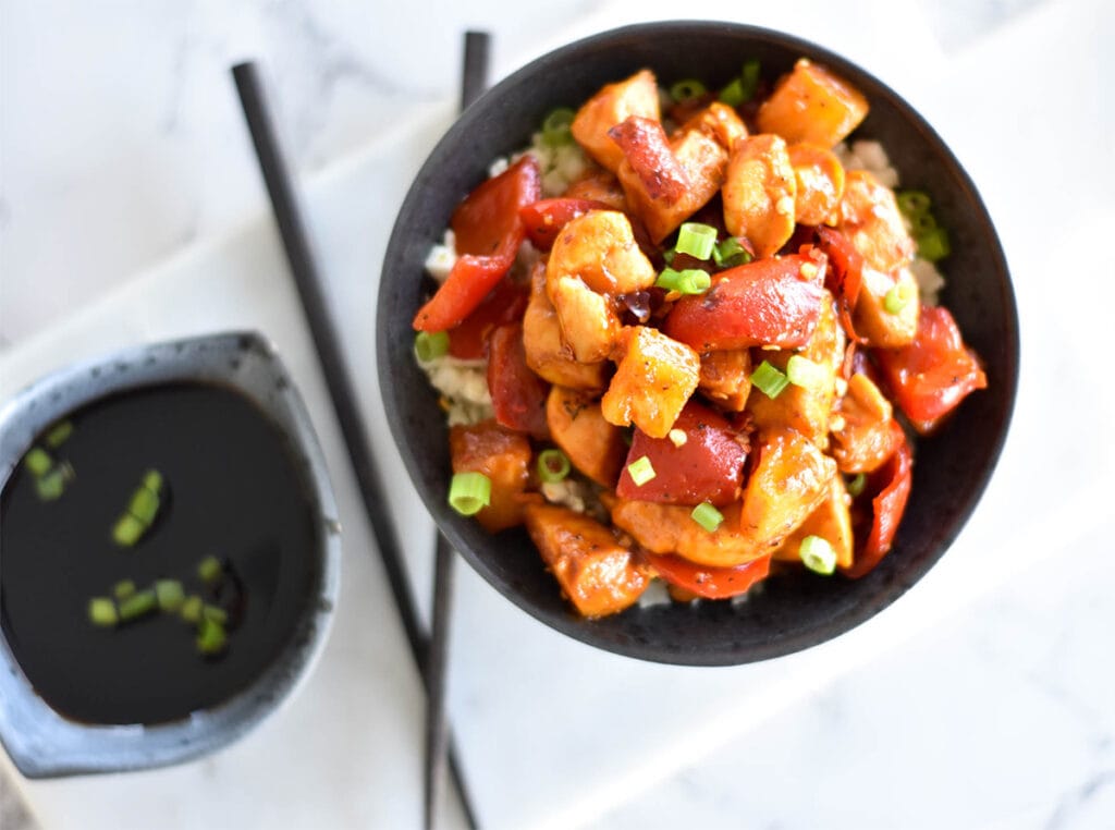 Top down view of chicken stir fry on bed of cauliflower rice with green onions next to a bowl of soy sauce.