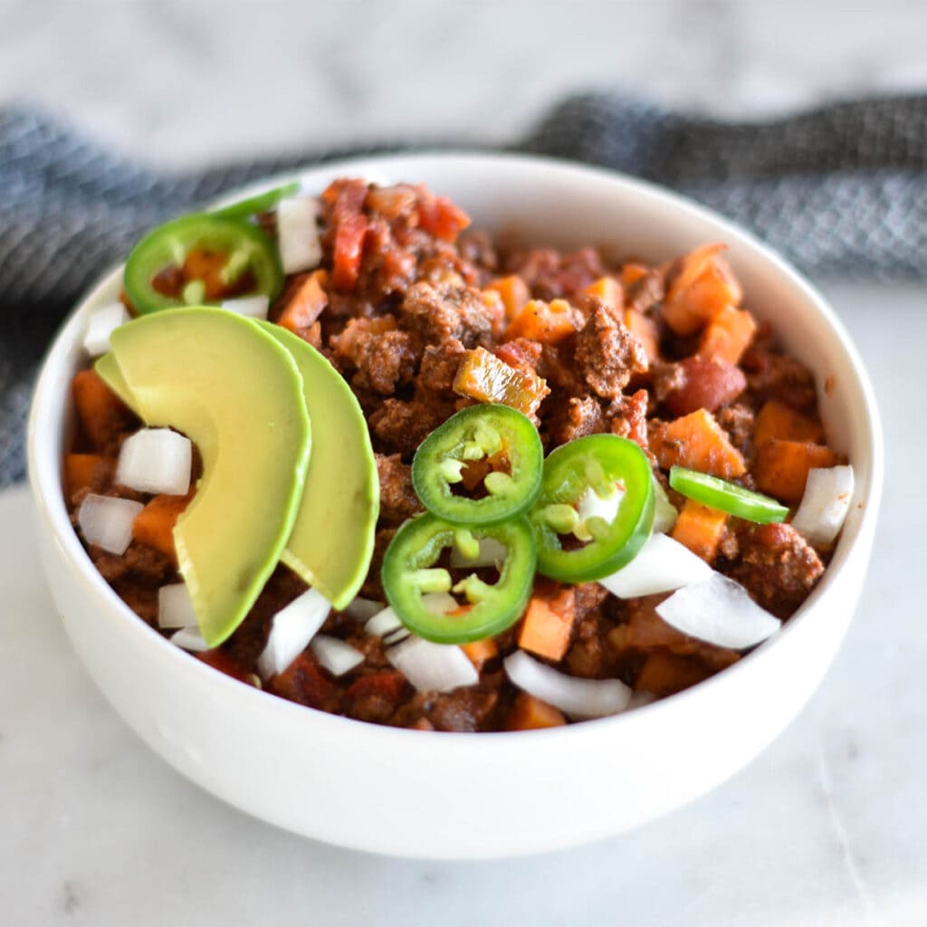 White bowl filled with sweet potato chili topped with avocado and jalapenos.