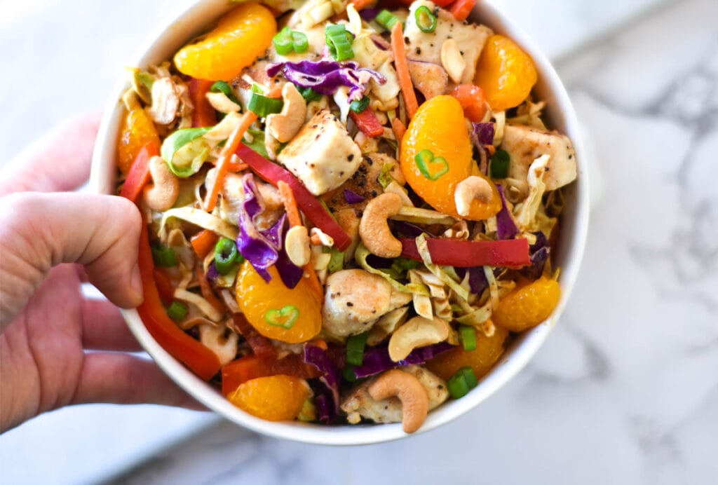 Hand holding a bowl of Whole30 Asian Chicken Salad with brightly colored mandarin oranges, bell peppers, green onions, cabbage and cashews.
