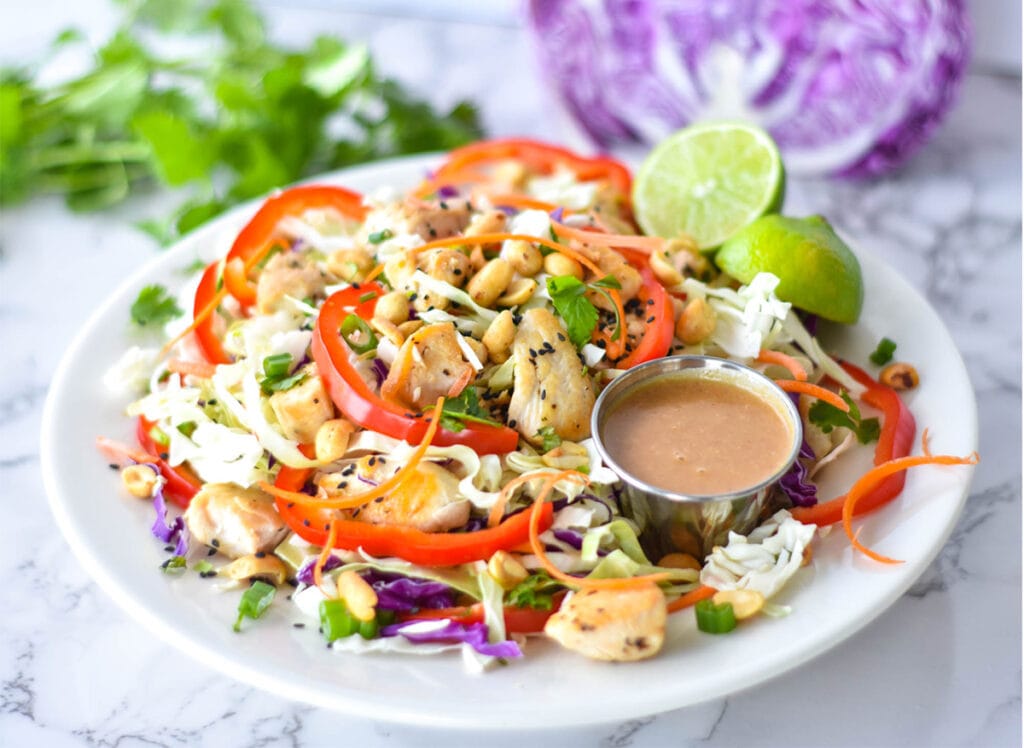 Large Thai chicken salad with red bell pepper, green and purple cabbage, chicken, peanuts and carrots next to a lime and peanut dressing.