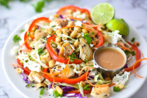 Large Thai chicken salad with red bell pepper, green and purple cabbage, chicken, peanuts and carrots next to a lime and peanut dressing.