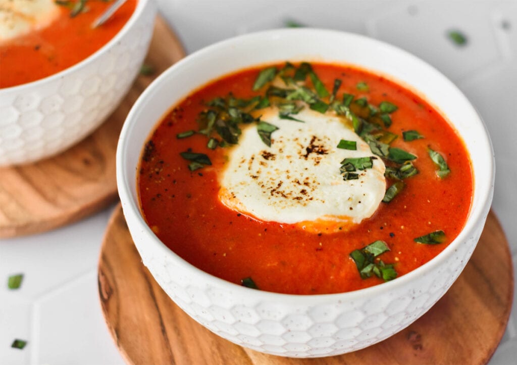 Bowl of bright red tomato soup topped with basil and melty cheese.