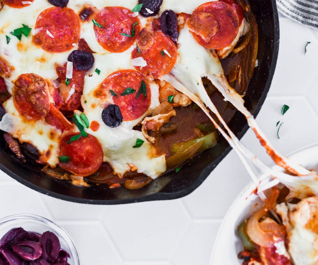 Cast iron skillet filled with keto pizza chicken skillet meal with melted cheese getting pulled out of the pan.