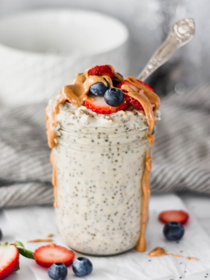 Mason jar overfilled with protein overnight oats topped with strawberries, blue berries and dripping peanut butter.