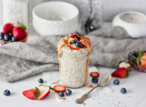 Mason jar overfilled with creamy protein powder overnight oats topped with strawberries, blue berries and dripping peanut butter.