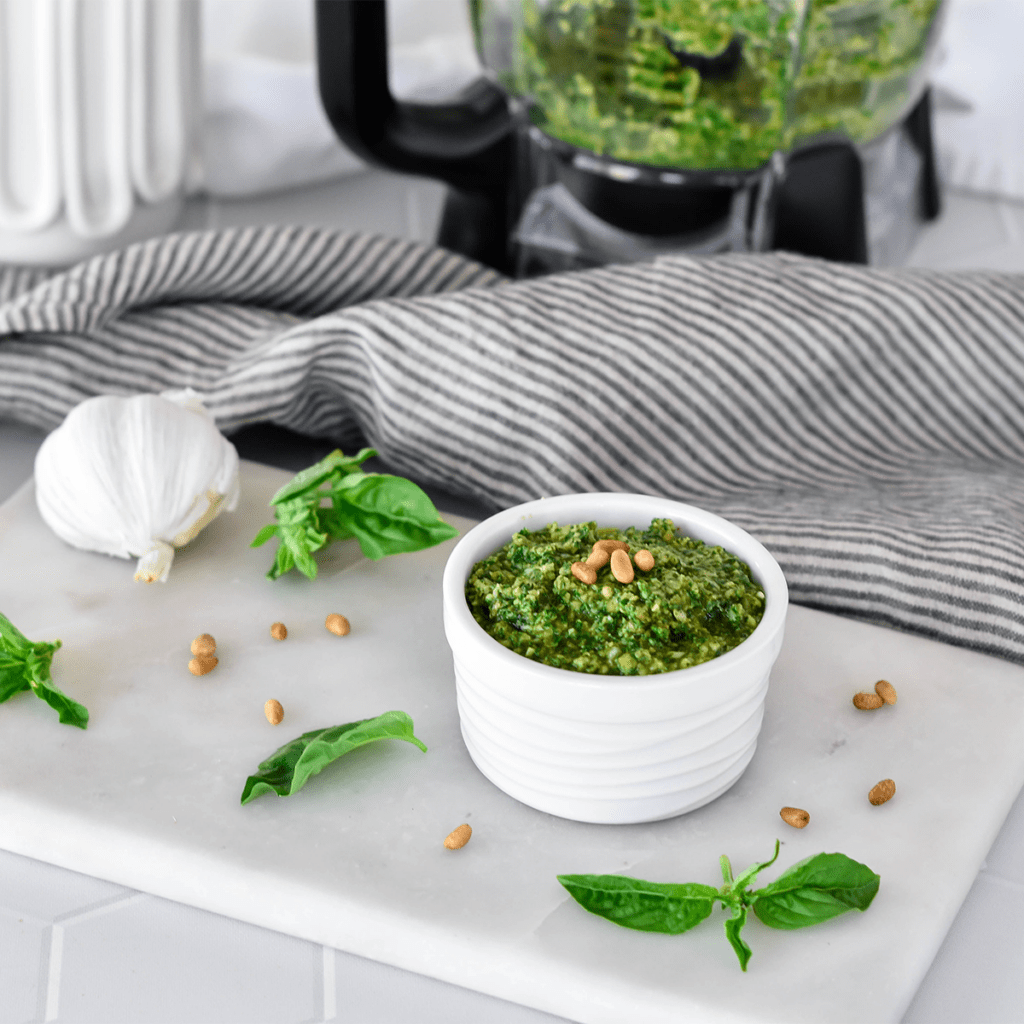 Dish filled with bright green pesto next to basil and garlic, topped with pine nuts.