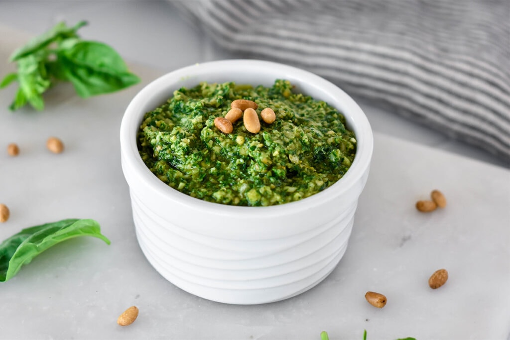 Dish filled with bright green dairy-free whole30 pesto next to basil and garlic, topped with pine nuts.