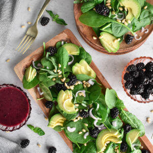 Wooden platter filled with greens, blackberries, avocado, pine nuts and shallot.