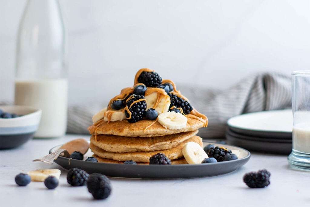 Stack of vegan oat pancakes with blackberries, peanut butter and banana slices on top.