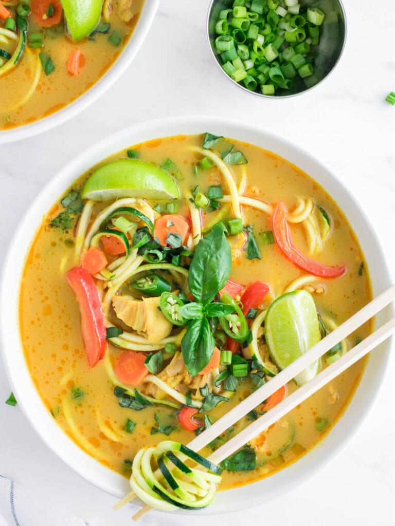 Paleo and Keto Thai Curry Chicken Zoodle Soup with basil, Thai chilis, bell pepper, lime, carrots, and chicken thighs.