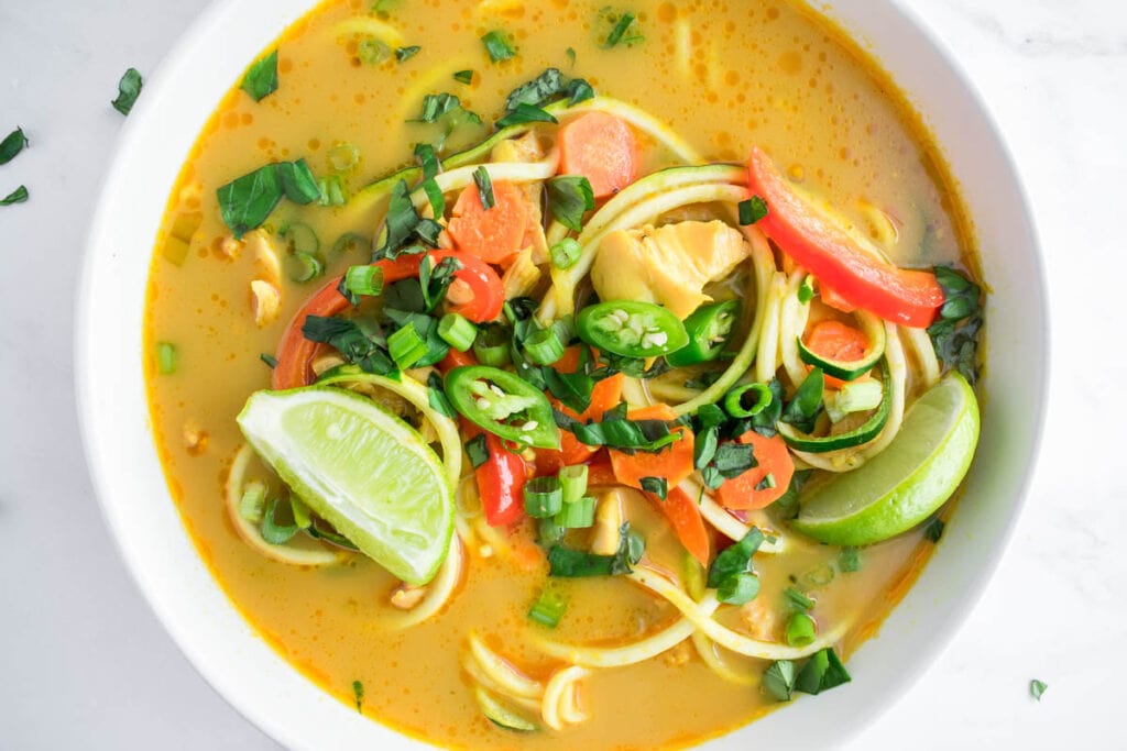 Paleo and Keto Thai Curry Chicken Zoodle Soup with basil, Thai chilis, bell pepper, lime, carrots, and chicken thighs.