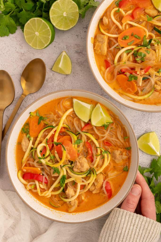 Red curry chicken soup garnished with limes and cilantro in a bowl with a hand holding the warm bowl.