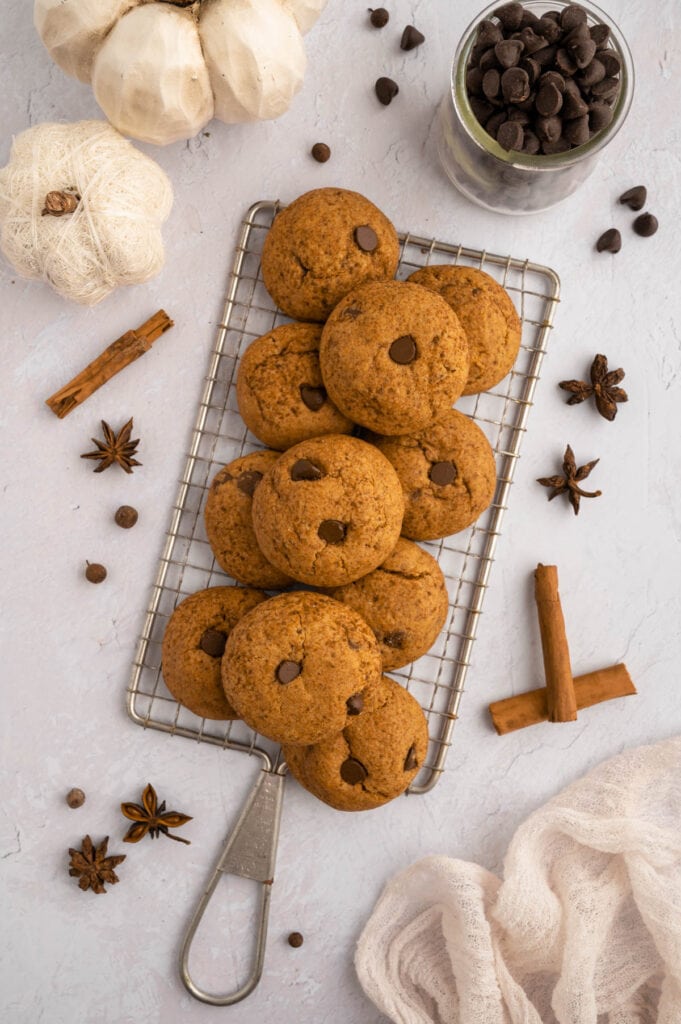 Wire rack of gluten free pumpkin chocolate chip cookies next to a bowl of chocolaye chips and cinnamon sticks..