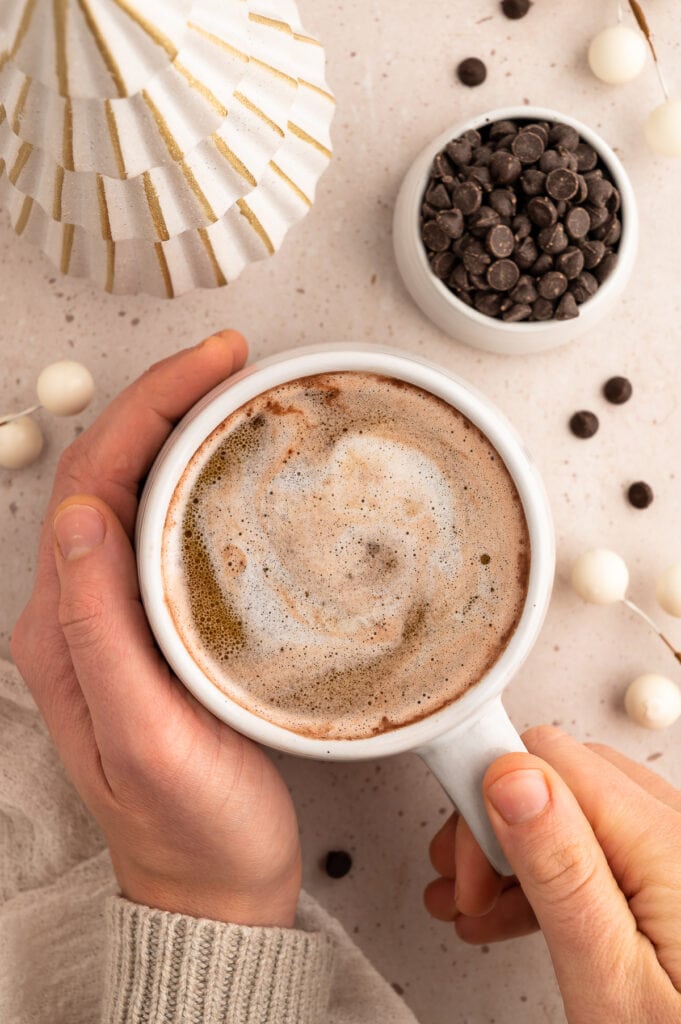 Hands holding a cup of keto hot chocolate with a ceramic christmas tree and a bowl of chocolate chips in the frame.