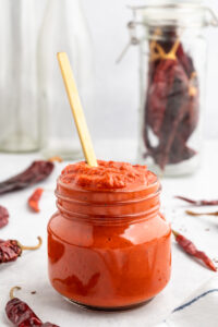 Jar of harissa sauce with a spoon in it next to peppers.